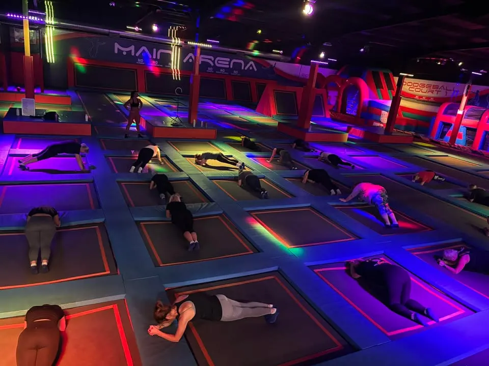 Multiple visitors bouncing and exercising on neon-lit individual trampolines in a brightly coloured indoor trampoline park.