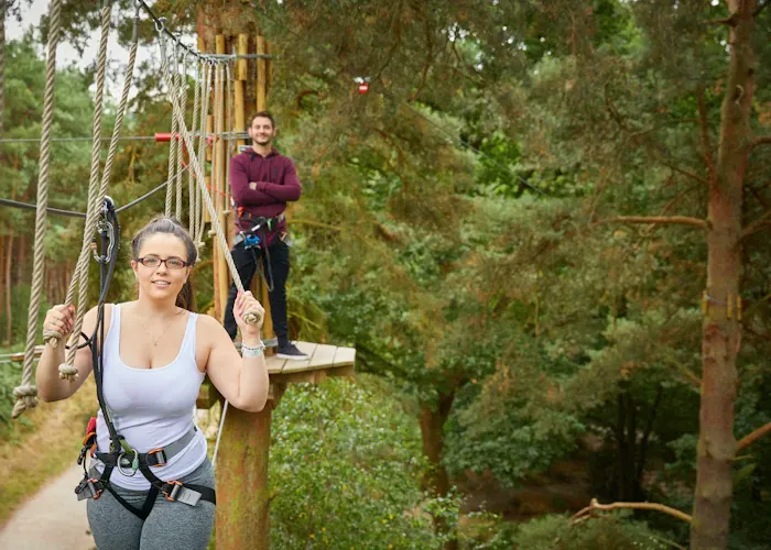 Woman in harness on forest canopy course with instructor watching from wooden platform.