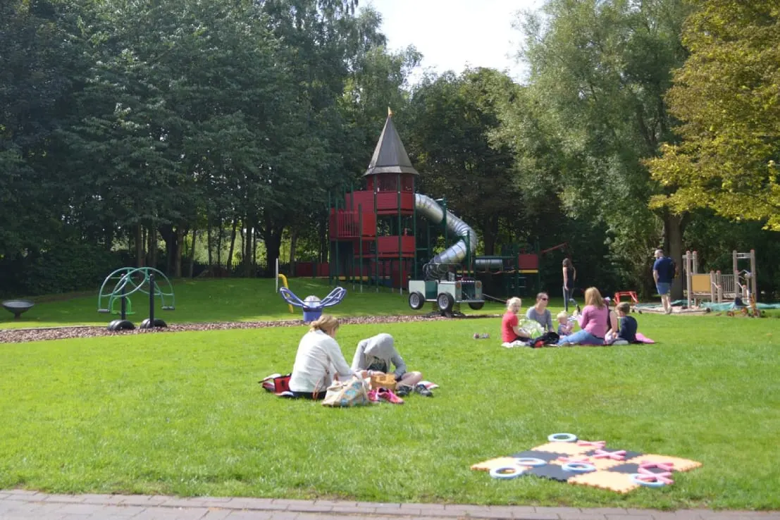 Children and adults enjoying an outdoor playground with a red castle-themed climbing structure, slide, and various play equipment on a grassy lawn surrounded by trees.