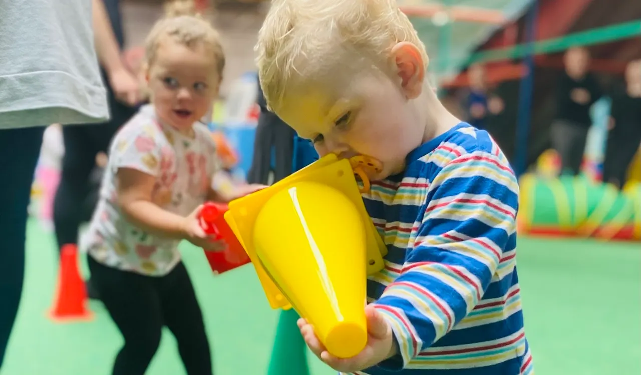 Young toddler in striped top holding a yellow foam toy whilst playing indoors with other children.