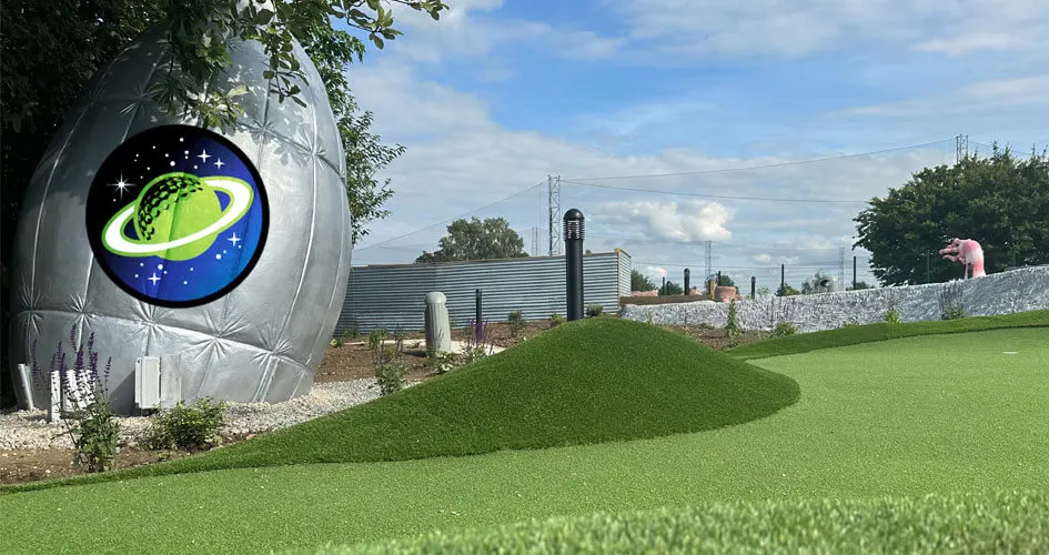Futuristic themed mini golf course with artificial turf, a large illuminated planet structure featuring a green ringed planet logo, and white barriers lining the fairway.