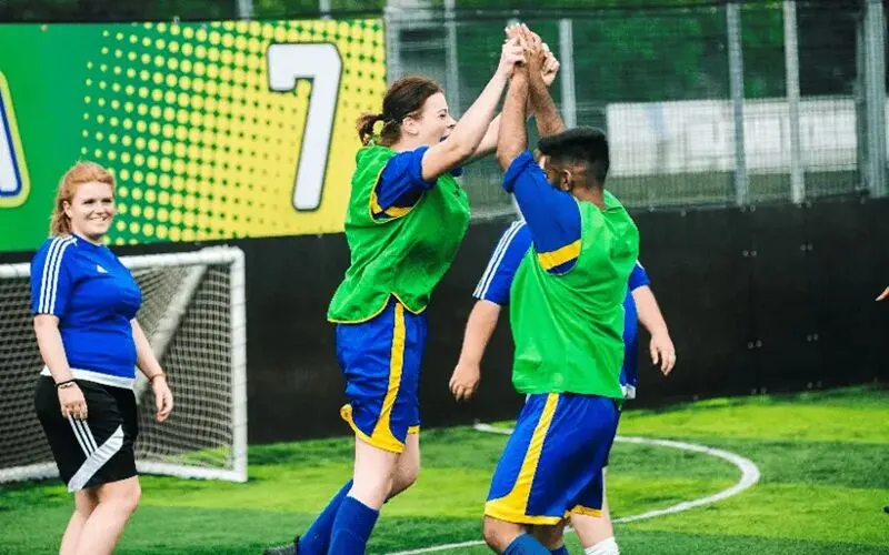 Young football players in green and blue kit celebrating with a high-five on an indoor pitch.