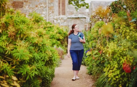 Woman jogging through a narrow garden pathway lined with flowering shrubs and climbing ivy-covered stone walls.