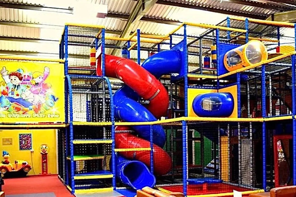 Multi-level indoor play structure with slides, tunnels and climbing frames in bright blue and red, housed in a warehouse setting.