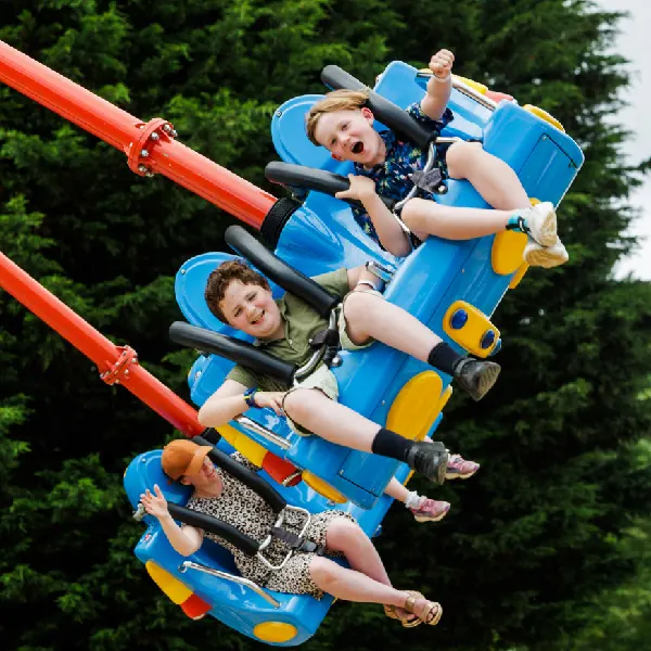 Three children ride a blue and yellow fairground ride with red track, expressions of excitement and joy on their faces.