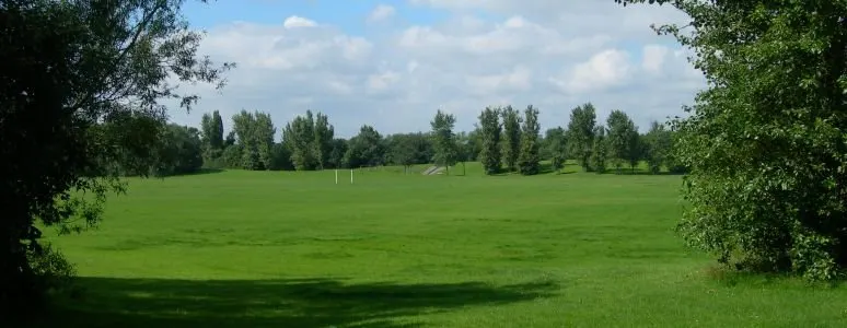 Expansive green sports field with trees lining the perimeter under a partly cloudy sky.