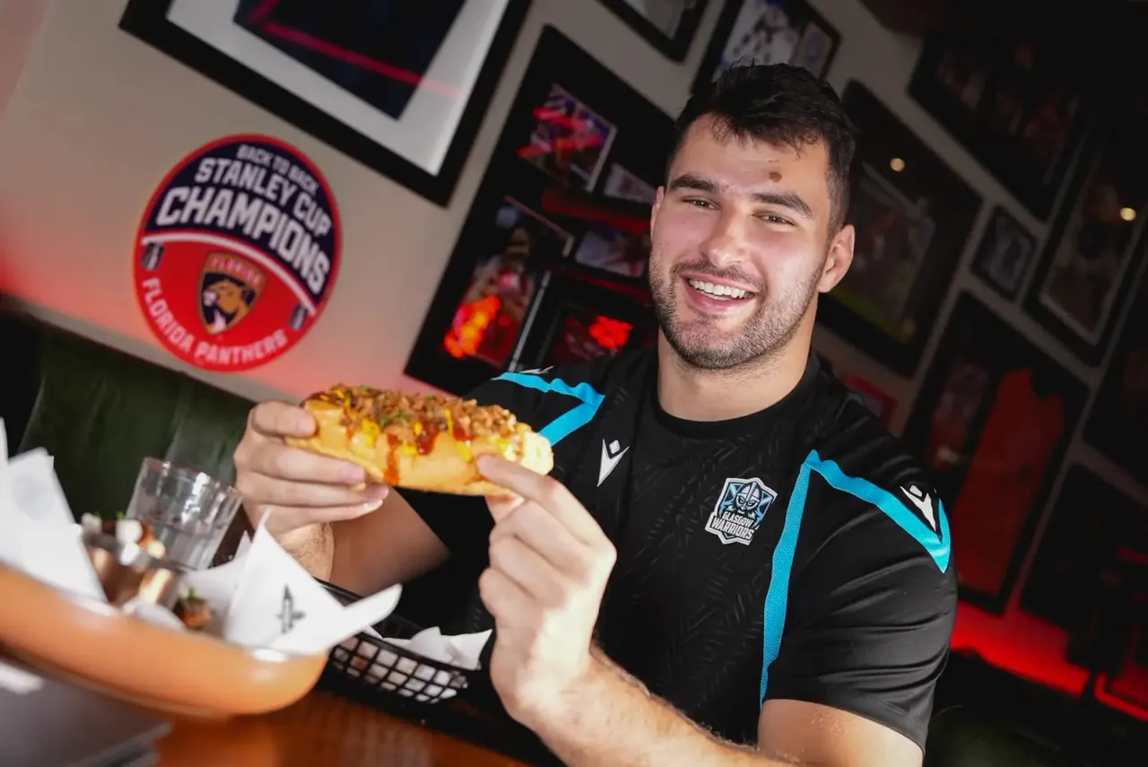Man in sports shirt smiling whilst holding a slice of pizza at an indoor venue with Stanley Cup Champions signage.