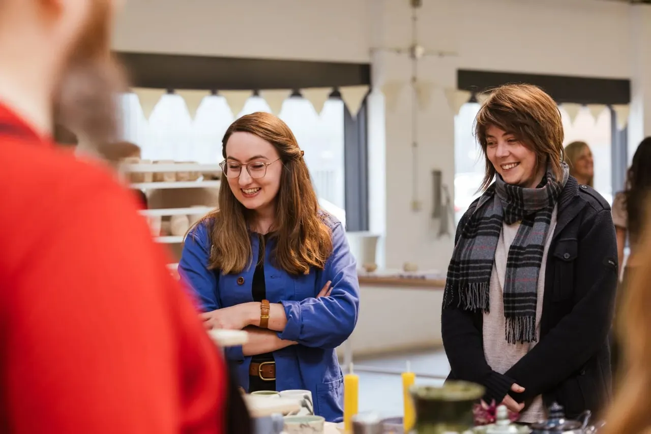 Two young women smiling whilst talking to someone at an indoor event with refreshments and bunting visible.