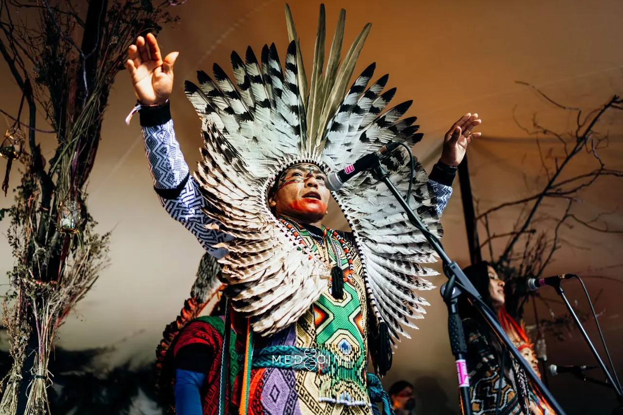 A performer wearing traditional indigenous regalia with a large feathered headdress sings passionately at a microphone on stage.