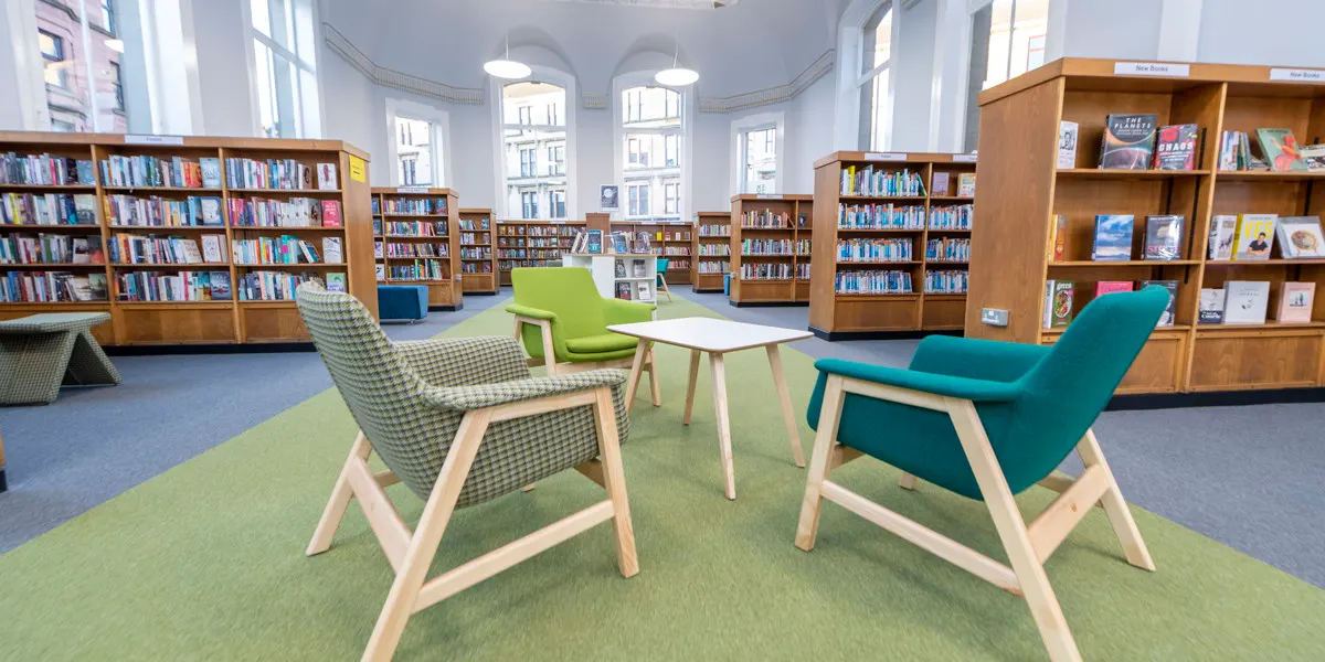 Modern library reading area with comfortable seating around a small table, surrounded by wooden bookshelves.