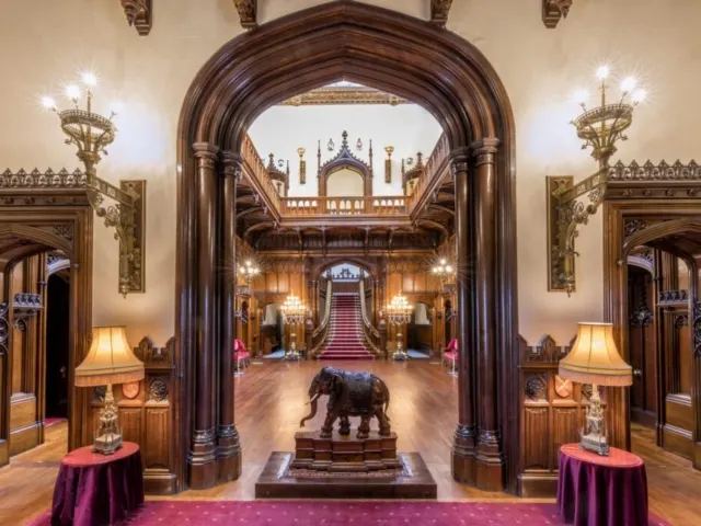 Grand ornate interior hallway with wooden archway, elephant statue, red carpet, and ornate staircase visible beyond.