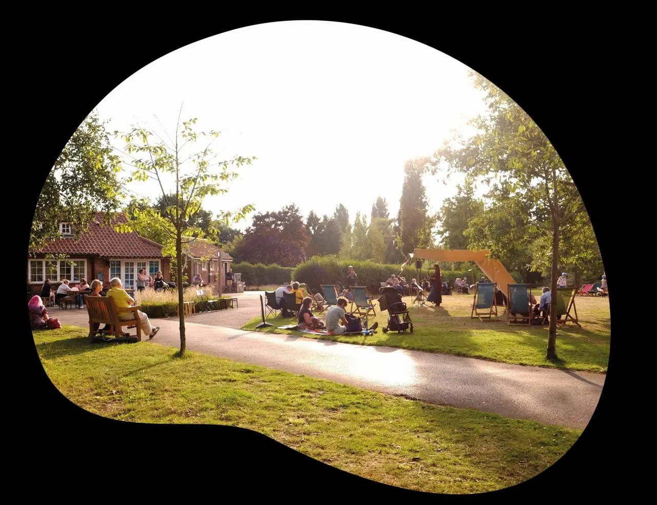 Visitors relaxing in a spacious parkland with brick buildings, pathways, seating areas and shaded structures on a sunny day.