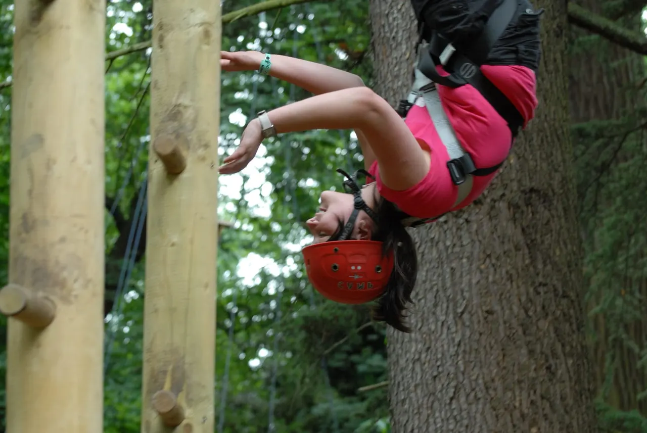 Child in pink harness and red helmet climbing wooden logs on an outdoor adventure course.