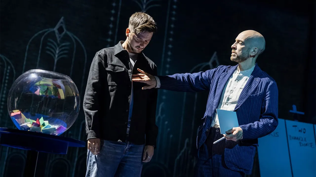 Two men on stage interact near a large illuminated sphere, with stylised botanical designs projected on the dark background behind them.