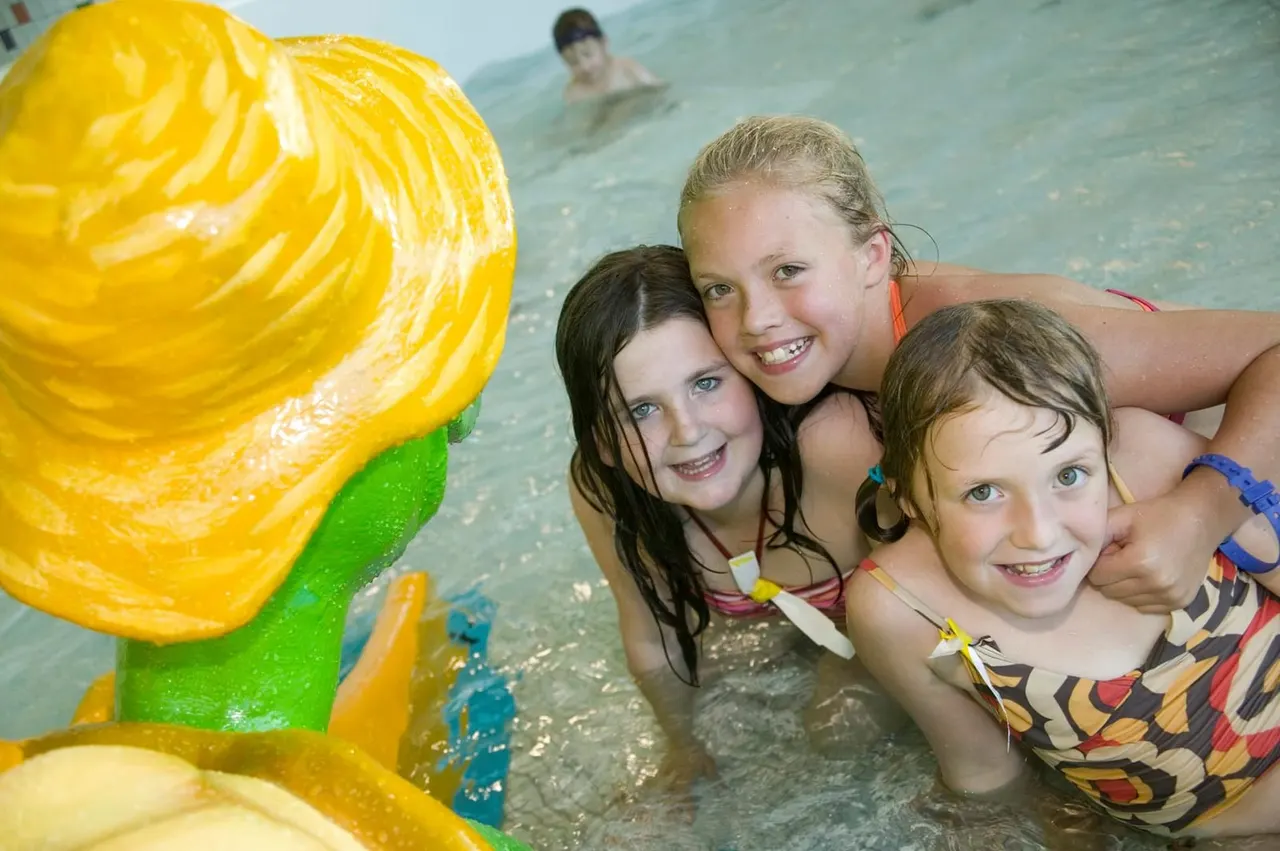 Three children playing in a shallow indoor pool next to a colourful yellow and green water toy.