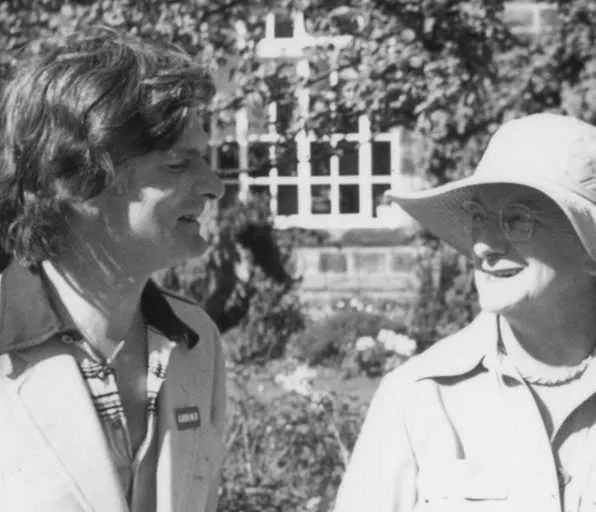 Two people converse in a garden with ivy-covered walls and a white lattice structure visible behind them.