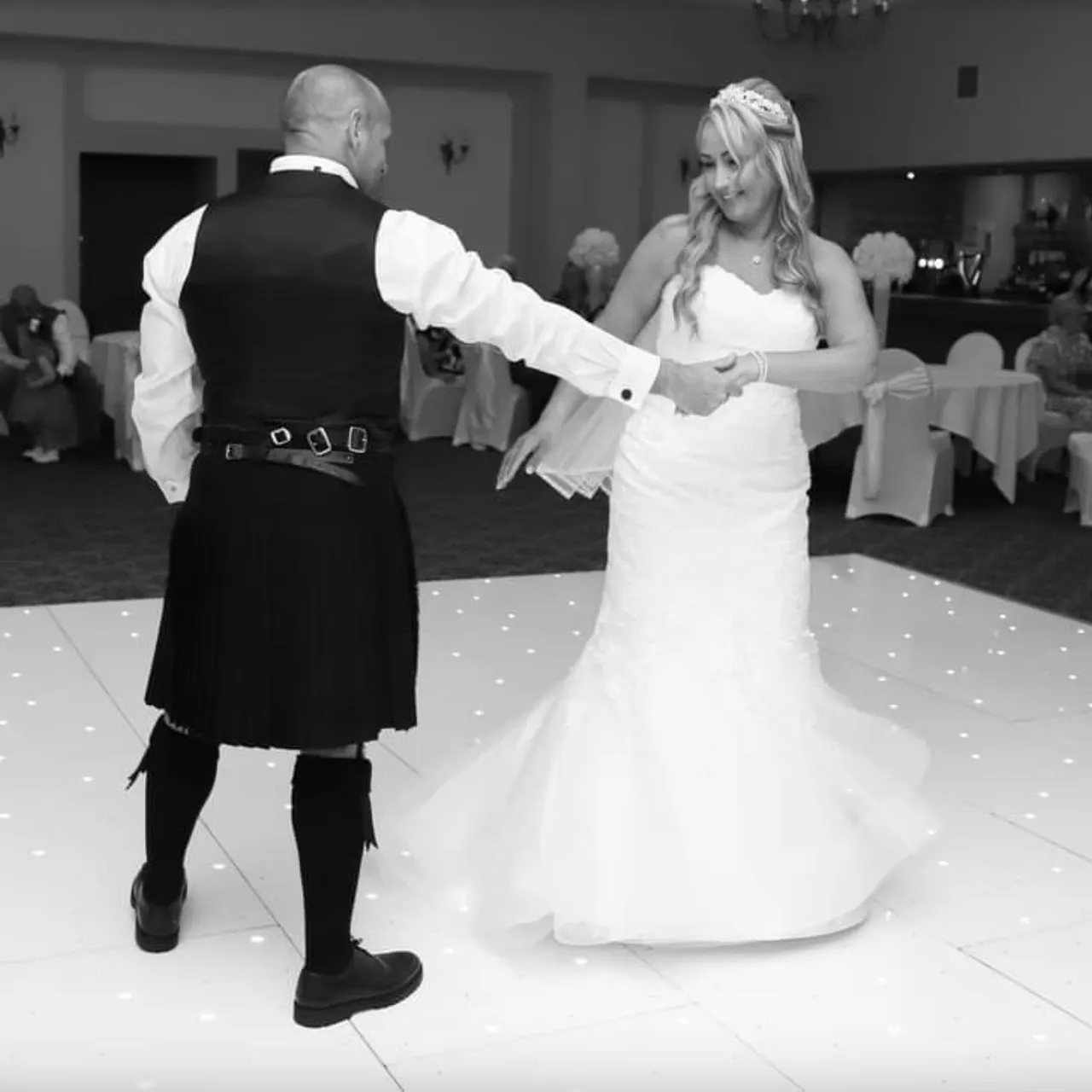Bride and groom dancing together on a lit dancefloor at a wedding reception, guests seated at tables in background.