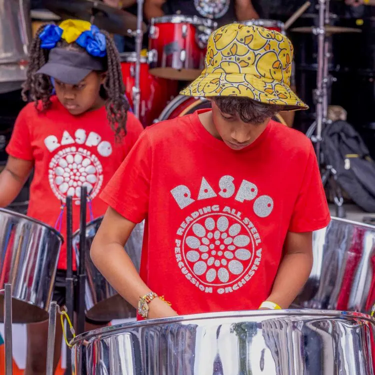 Two children in red branded t-shirts and colourful hats playing steel drums at an outdoor music event.