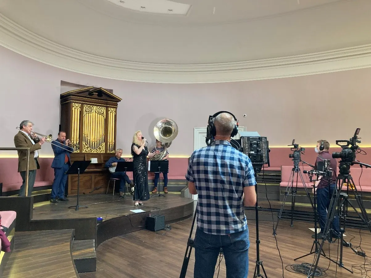 Musicians performing with brass instruments on stage in concert hall whilst being filmed by camera crew.