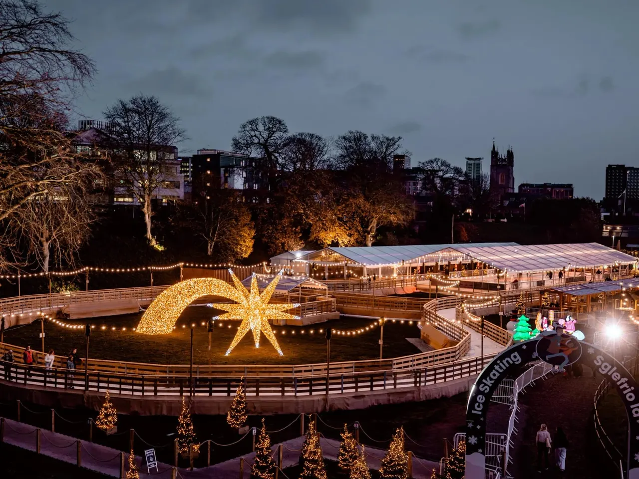 Illuminated outdoor winter ice skating venue with large golden star sculpture, decorated trees, marquees, and city skyline at dusk.