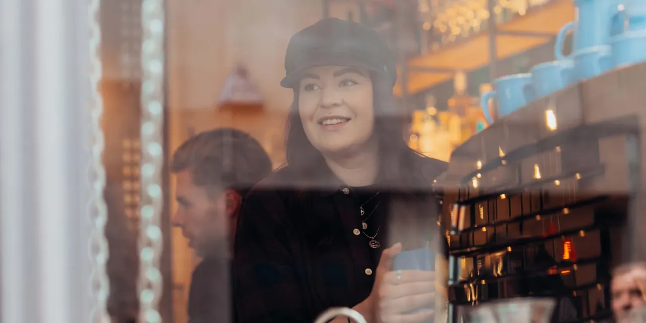 Woman in black cap and coat smiling whilst looking out through a shop window at an illuminated urban street at dusk.