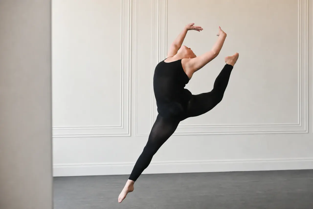 Dancer in black clothing performing a leaping jump with arms raised against a white panelled wall.