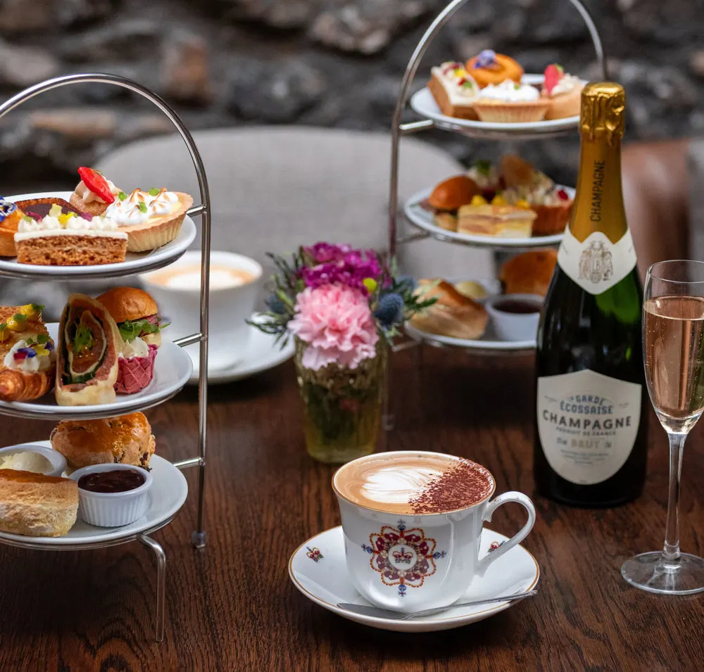 Tiered afternoon tea spread with sandwiches, pastries, cakes, champagne and cappuccino on wooden table.