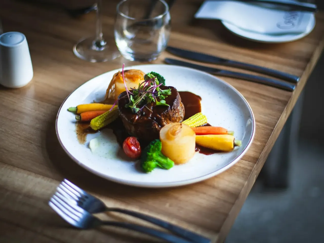 Plated fine dining dish with glazed meat, roasted vegetables and microgreens on wooden table setting.