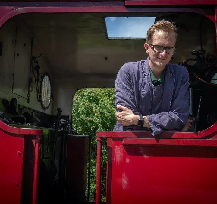 Man in glasses and purple jacket stands in the cab of a red steam locomotive.