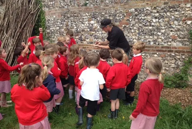 An adult instructor demonstrates something to a group of children wearing red clothing gathered outdoors against a stone wall.