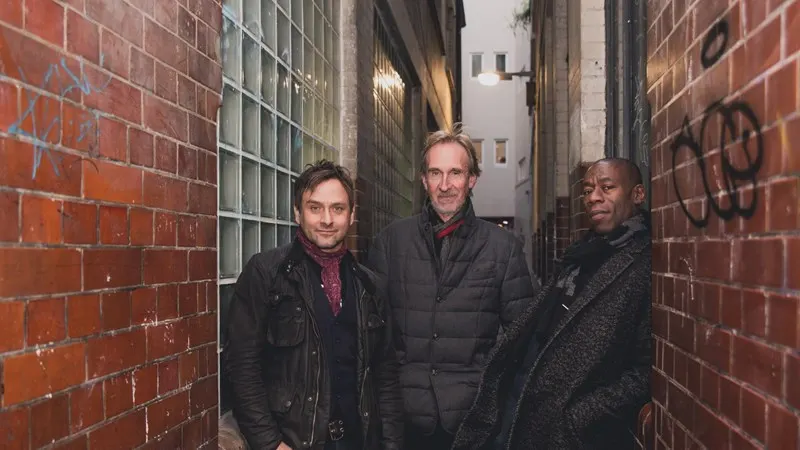 Three men standing in a narrow urban alleyway between brick buildings.