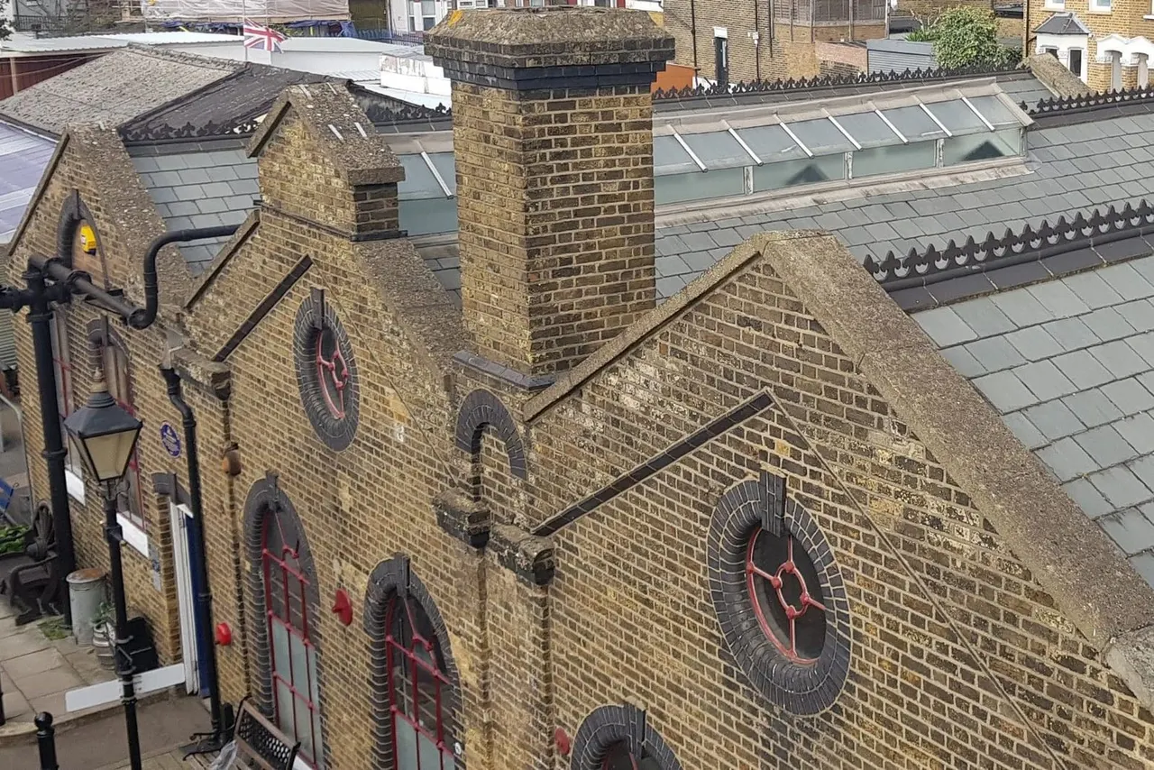 Aerial view of historic brick buildings with decorative clock faces and red doors, surrounded by London rooftops and chimneys.