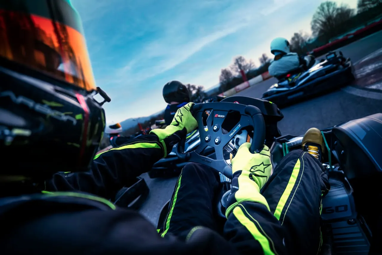 Multiple go-kart drivers in neon racing suits and helmets competing on an outdoor track under blue sky.