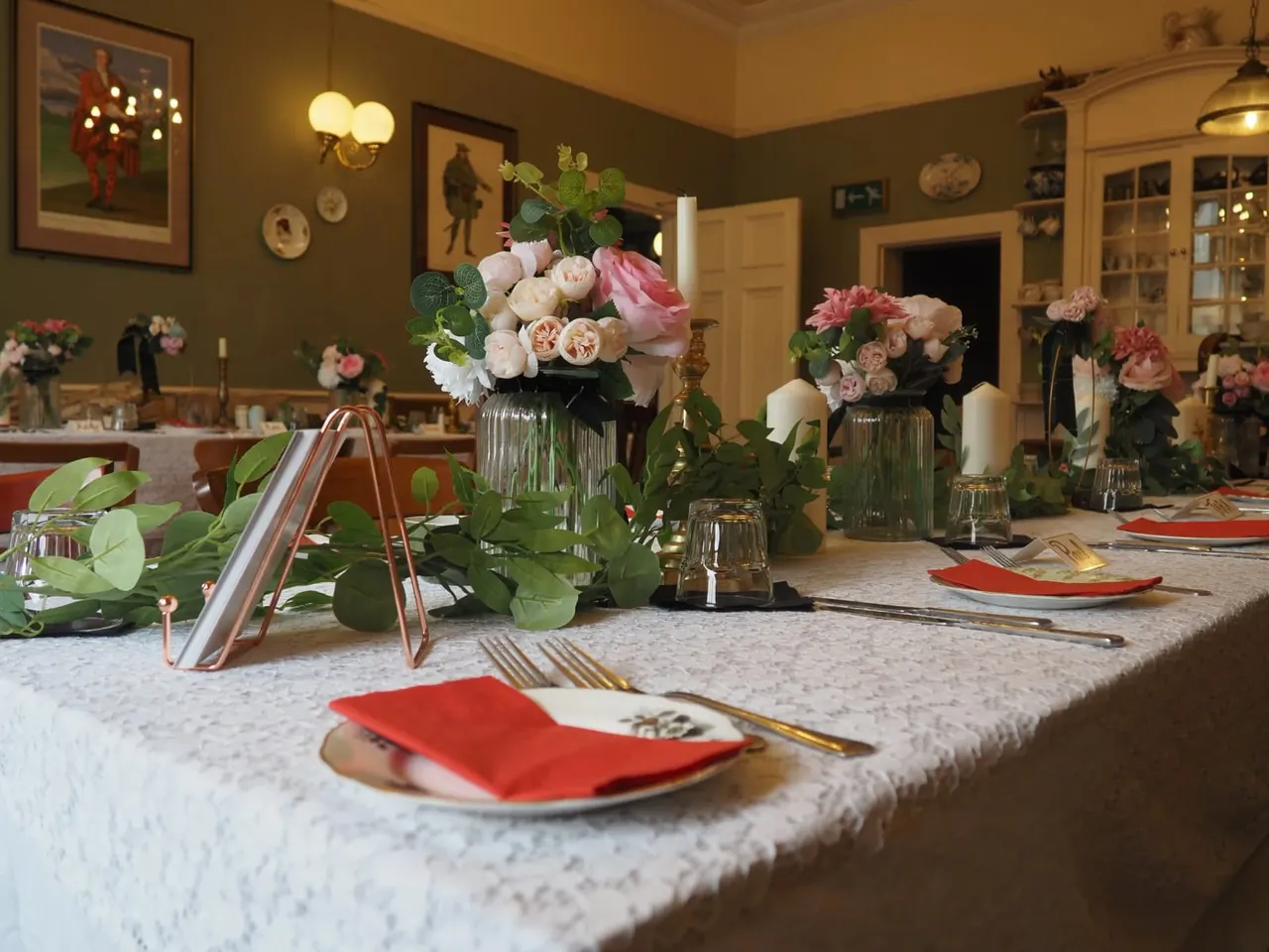 Elegant dining table set for an event with pink and white floral arrangements in glass vases, red napkins, and gold cutlery in a decorated room.