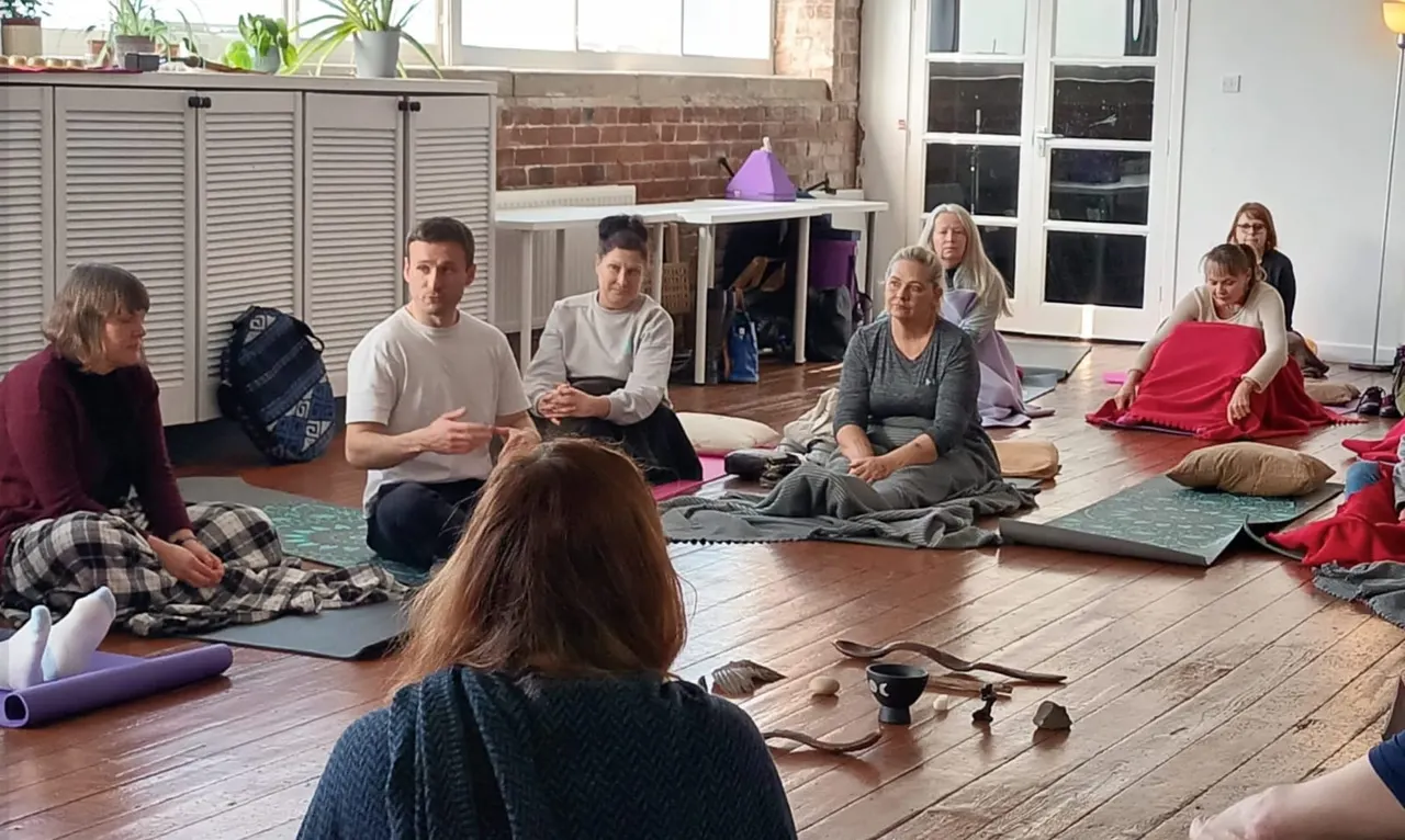 A group of adults sitting on yoga mats in a circle on a wooden studio floor during a mindfulness or meditation session.