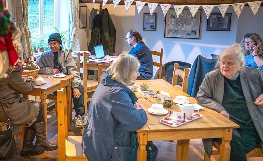 A group of older adults socialise over refreshments at wooden tables in a decorated community room with bunting and framed pictures on the walls.