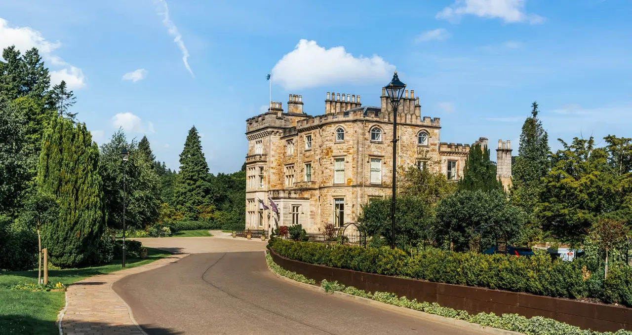 Stone castle with crenellated towers and manicured grounds, approached via curved driveway lined with trees and hedges under blue sky.