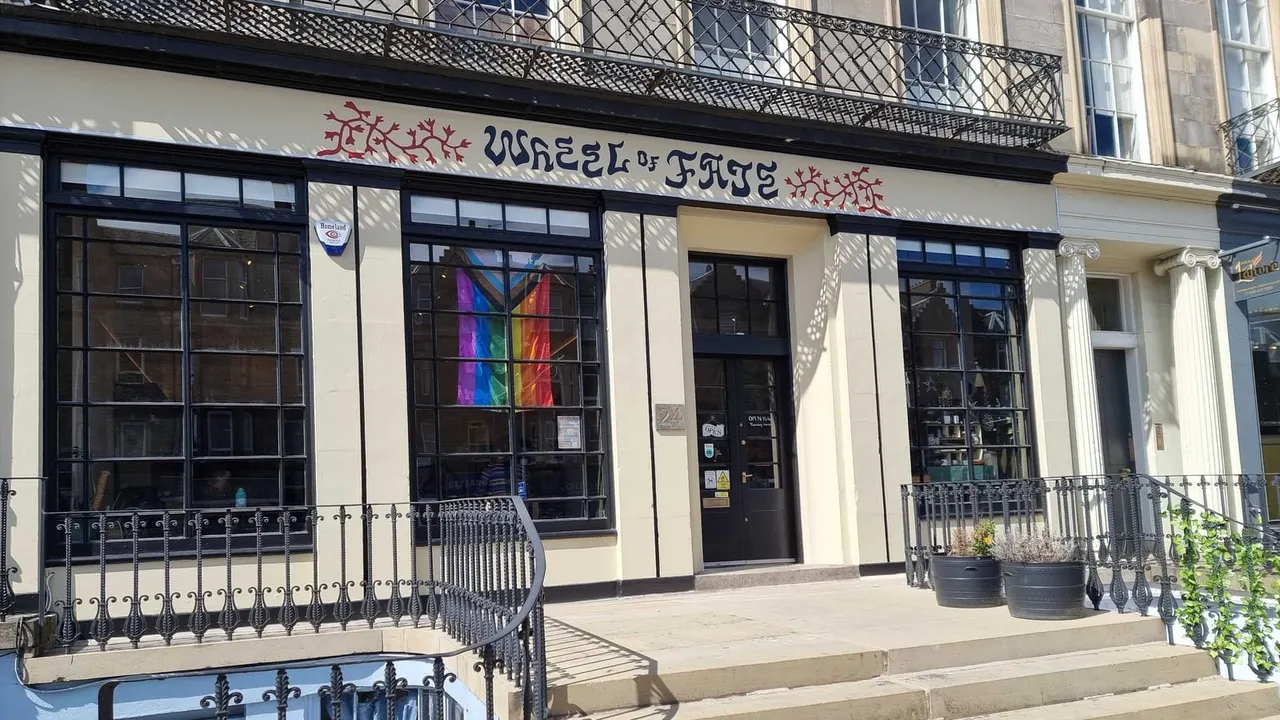 Storefront of Wheel of Fate venue with cream and black façade, large windows, rainbow flag, and metal railings.