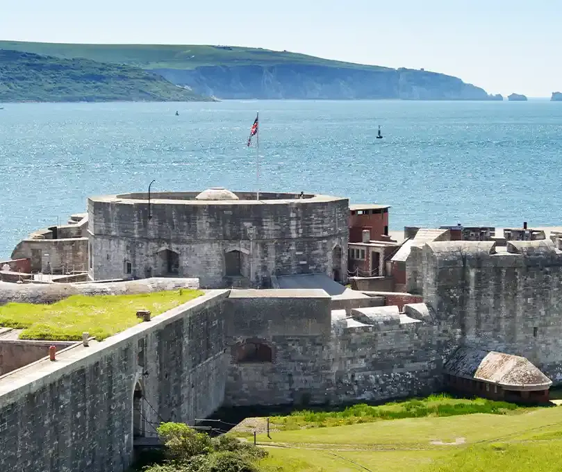 Historic stone fortress with flag flying, green grass surroundings, sea and white cliffs visible beyond.