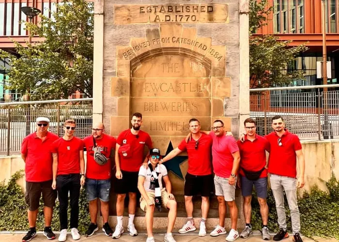 A group of eight people wearing matching red polo shirts stand in front of a stone brewery building established in 1770.