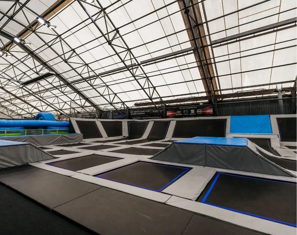 Indoor trampoline park with interconnected black and blue trampolines beneath an industrial glass and metal roof structure.