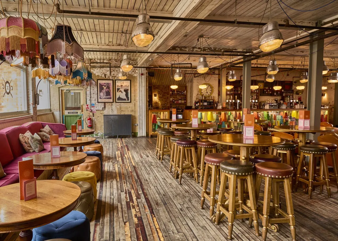 Spacious bar and lounge interior with wooden furniture, vintage pendant lighting, colourful seating, and shelves of bottles.