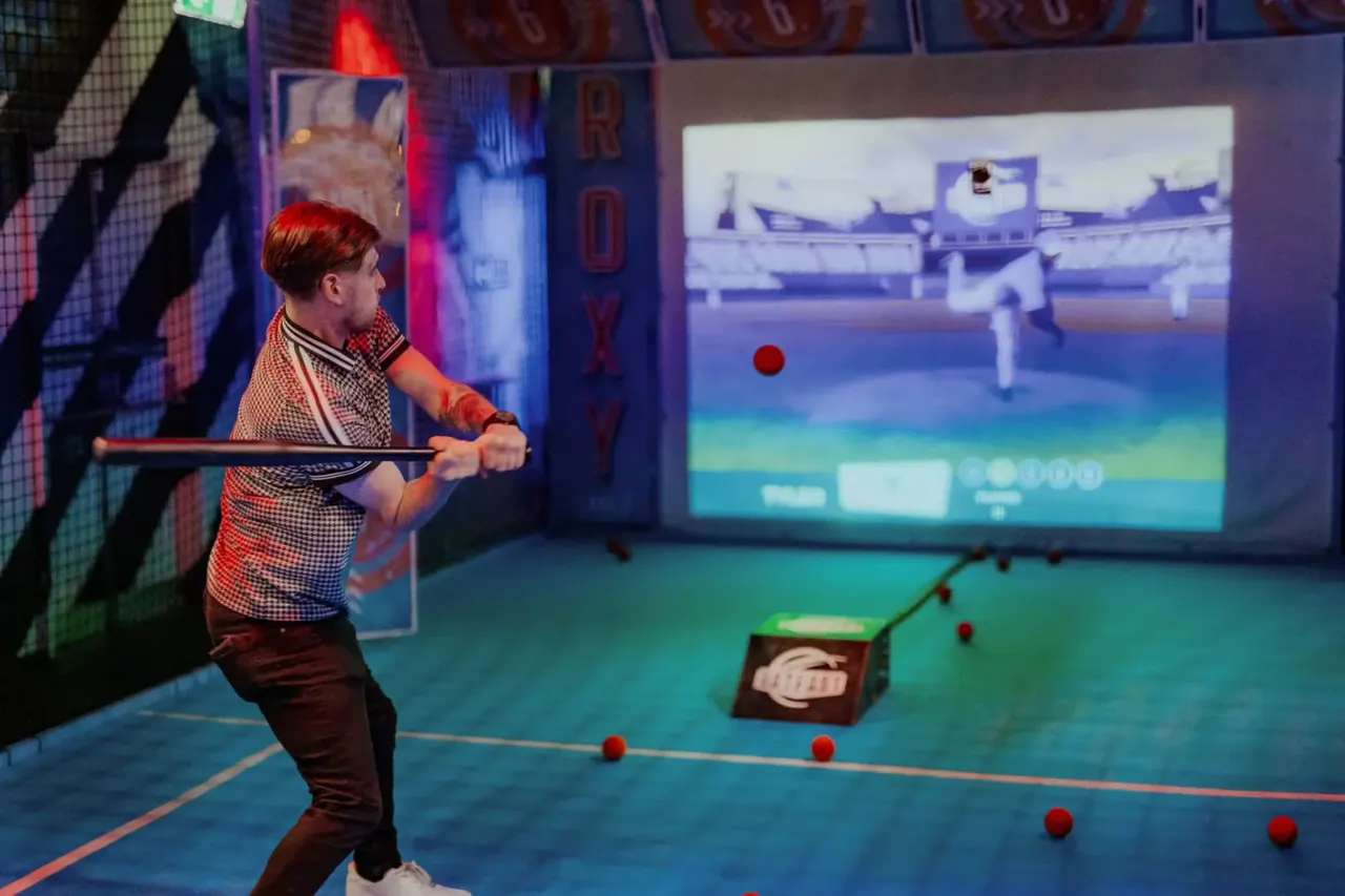 A young person playing an interactive baseball video game, swinging at a virtual ball projected on a screen in a brightly lit arcade game zone.