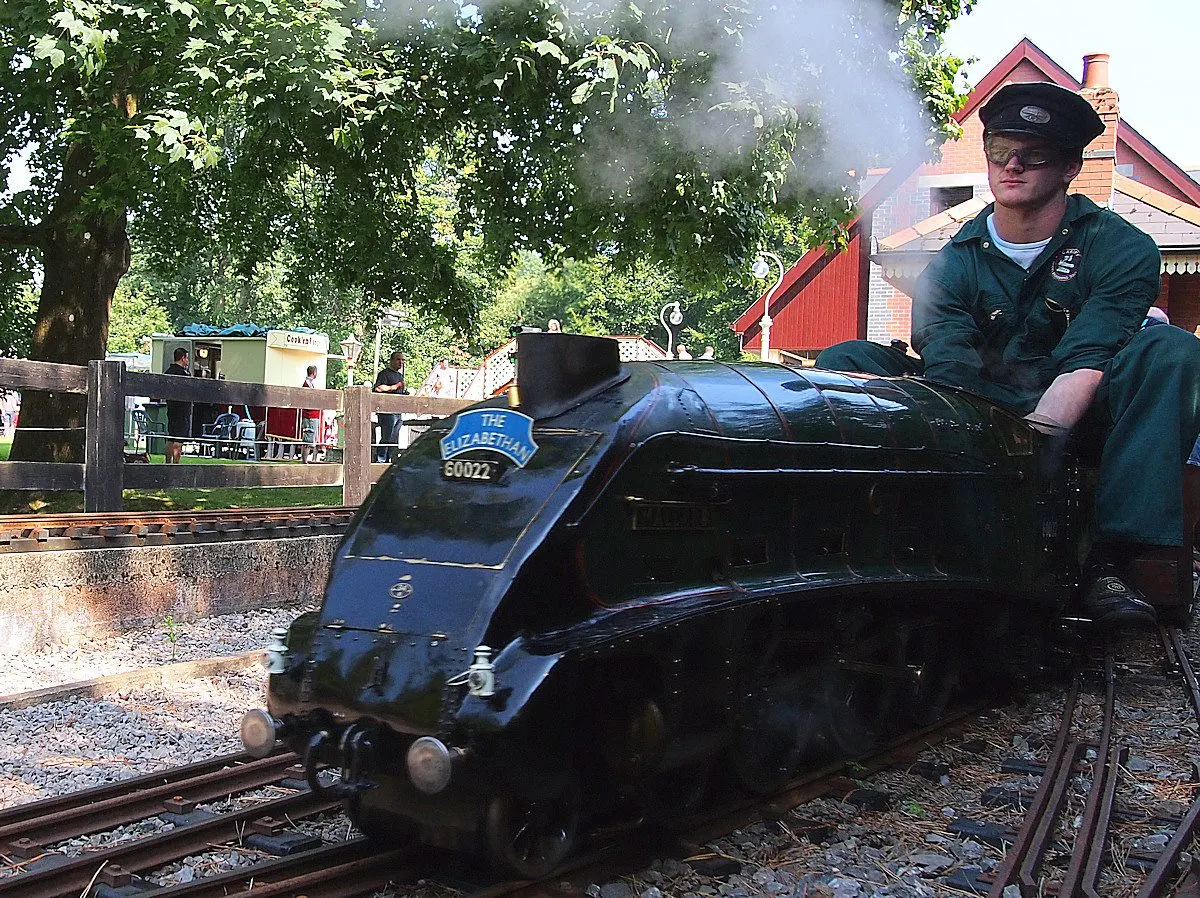 A man operates a miniature steam locomotive on narrow-gauge railway tracks, with a red barn and trees in the background.