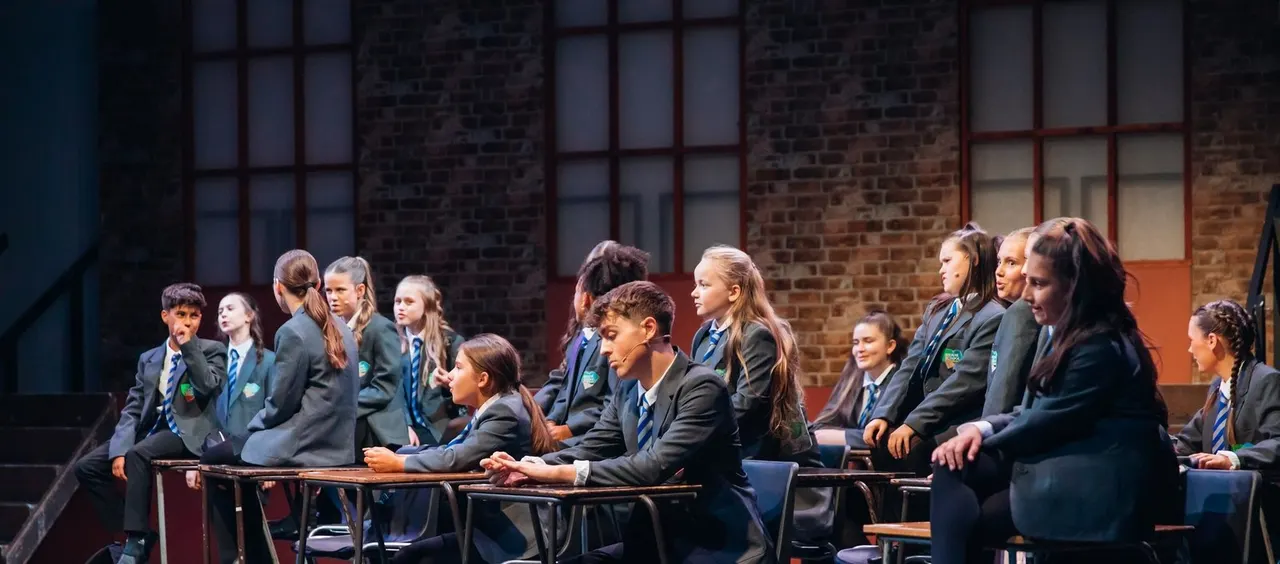 School students in uniform sitting together on a theatrical stage set with brick walls.