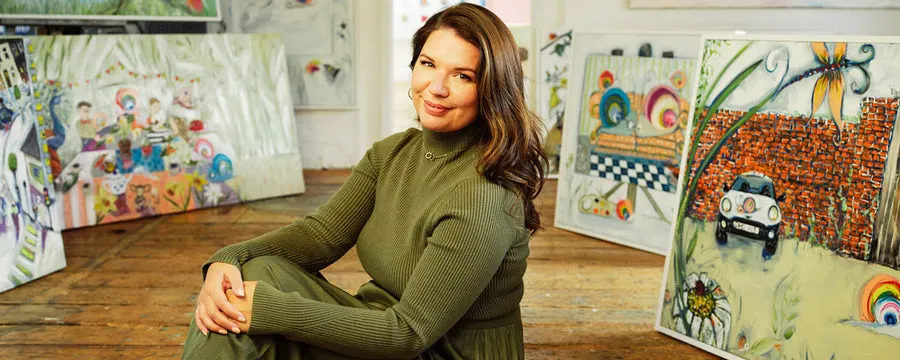 Woman posing in art studio surrounded by colourful artwork and paintings on wooden floor.