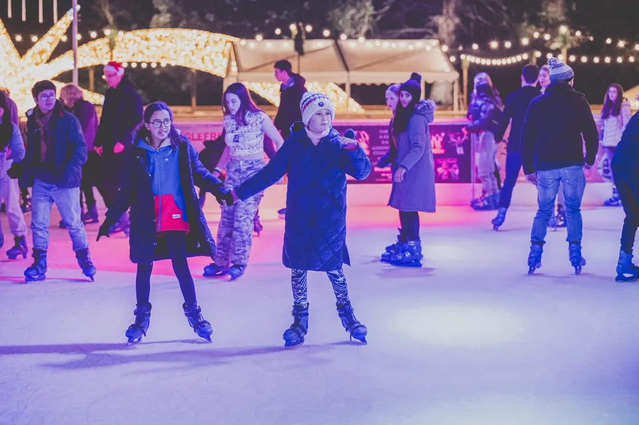 Groups of visitors ice skating at an outdoor rink lit by purple lighting and festive decorations.