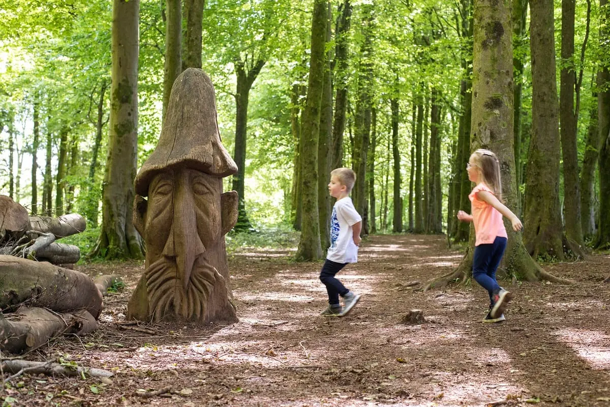 Two children running along a woodland path past a wooden carved figure and felled logs.