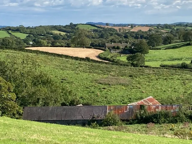 Rolling green pasture with disused farm wagons, fields and distant hills under blue sky.