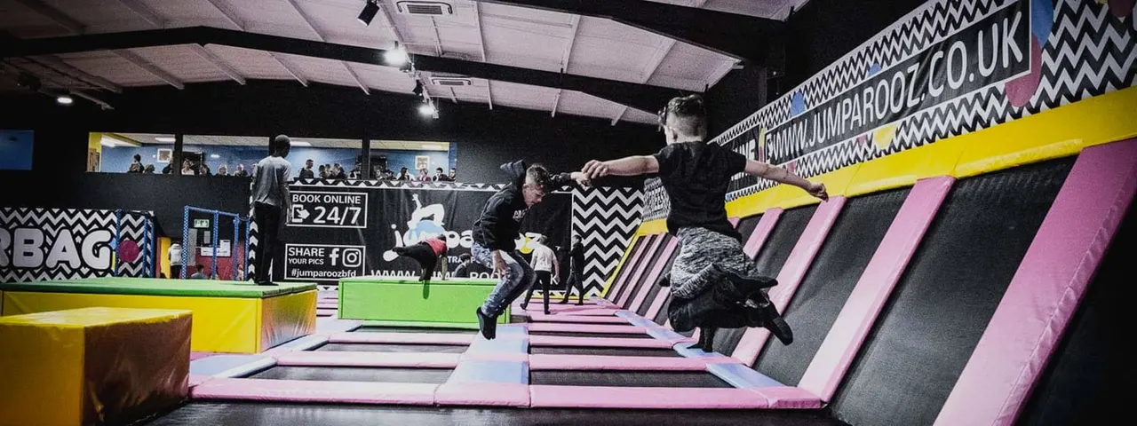 Children and adults bouncing and jumping on pink and grey interconnected trampolines inside a brightly coloured indoor play facility.